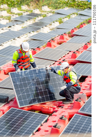 Vertical image of two technician workers help to set up or install solar cell panel into base over water reservoir. Vertical image of two technician workers help to set up or install solar cell panel into base over water reservoir. 111700386