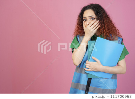 Front view of shocked, surprised woman with red, cturly hair standing, holding folders, closing mouth. Concept of aid. Front view of shocked, surprised woman with red, cturly hair standing, holding folders, closing mouth. Concept of aid. 111700968