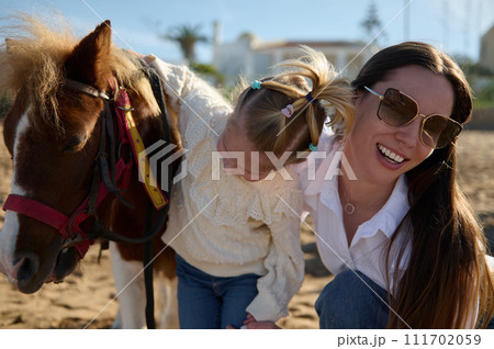 Happy young mother and daughter admiring together a pony horse. Family engaging in outdoors activities with little child Happy young mother and daughter admiring together a pony horse. Family engaging in outdoors activities with little child 111702059