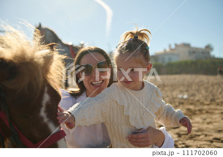 Happy mother carrying her daughter while stroking a little pony outdoor Happy mother carrying her daughter while stroking a little pony outdoor 111702060