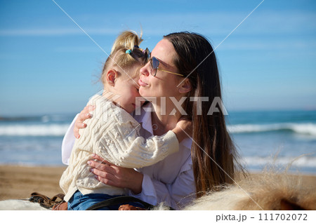 Happy affectionate mother cuddling her little daughter who learns riding a pony outdoor Happy affectionate mother cuddling her little daughter who learns riding a pony outdoor 111702072