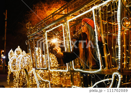 Woman holding sparkler night while celebrating Christmas outside. Dressed in a fur coat and a red headband. Blurred christmas decorations in the background. Selective focus 111702089