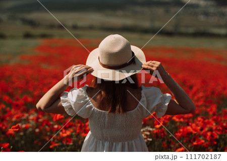 Field poppies woman. Happy woman in a white dress and hat stand through a blooming field of poppy raised her hands up. Field of blooming poppies. 111702187
