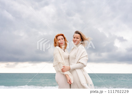 Women sea walk friendship spring. Two girlfriends, redhead and blonde, middle-aged walk along the sandy beach of the sea, dressed in white clothes. Against the backdrop of a cloudy sky and the winter Women sea walk friendship spring. Two girlfriends, redhead and blonde, middle-aged walk along the sandy beach of the sea, dressed in white clothes. Against the backdrop of a cloudy sky and the winter 111702229