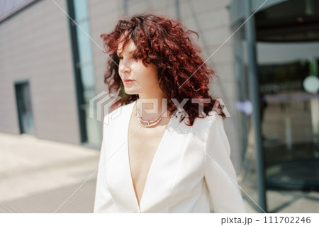 Portrait of a woman standing near a supermarket building. Caucasian model with long dark hair, wearing a white jacket and colored trousers. 111702246