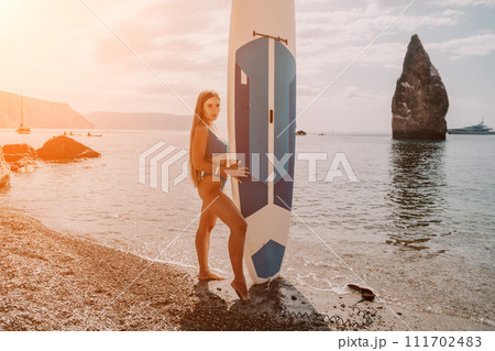 Woman sea sup. Close up portrait of happy young caucasian woman with long hair looking at camera and smiling. Cute woman portrait in bikini posing on sup board in the sea Woman sea sup. Close up portrait of happy young caucasian woman with long hair looking at camera and smiling. Cute woman portrait in bikini posing on sup board in the sea 111702483