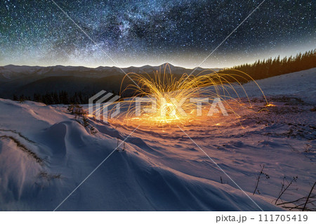 Light painting art. Spinning steel wool in abstract circle, firework of bright yellow glowing sparkles on snowy white valley on woody mountain ridge and blue night starry sky copy space background. 111705419