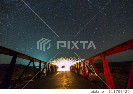 Light painting art concept. Long exposure shot of spinning steel wool in abstract circle making firework showers of bright yellow glowing sparkles on long bridge on blue night starry sky background. 111705420