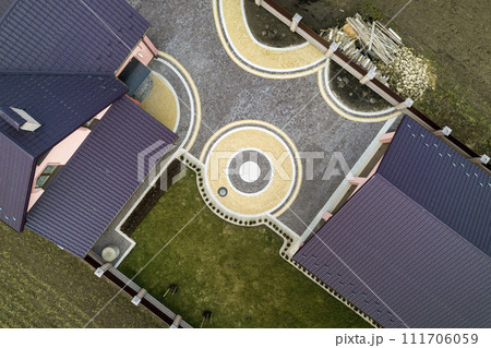 Aerial top view of house shingle roof on background of green lawn and colorful paved yard with geometrical abstract pattern. Roofing, repair and renovation work. Aerial top view of house shingle roof on background of green lawn and colorful paved yard with geometrical abstract pattern. Roofing, repair and renovation work. 111706059