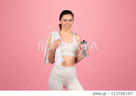 A happy, athletic woman in a white sports bra and leggings holds a water bottle and a towel over her shoulder A happy, athletic woman in a white sports bra and leggings holds a water bottle and a towel over her shoulder 111706187