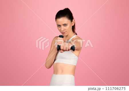 Focused young woman in white sportswear engaging in a strength training session with a black dumbbell Focused young woman in white sportswear engaging in a strength training session with a black dumbbell 111706275