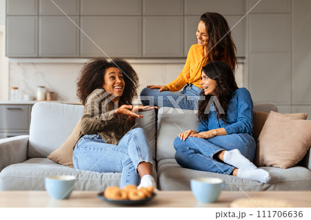 Multicultural trio of ladies chats joyfully around living room table 111706636