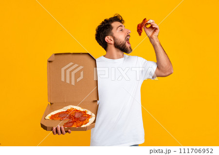 guy holding delivery box and savoring pizza slice, yellow backdrop guy holding delivery box and savoring pizza slice, yellow backdrop 111706952
