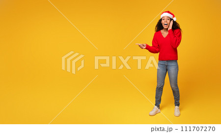Surprised black lady in Santa hat points at space, yellow background Surprised black lady in Santa hat points at space, yellow background 111707270
