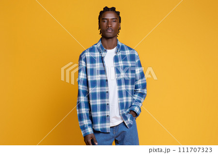 Stylish young black man wearing blue checkered shirt posing on yellow background 111707323