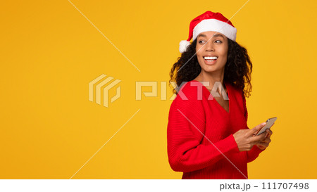 Black lady with cellphone in hands, woman wearing Santa hat, looking back at free space 111707498