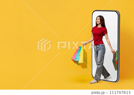 Cheerful young asian lady with purchases walking from huge phone Cheerful young asian lady with purchases walking from huge phone 111707613