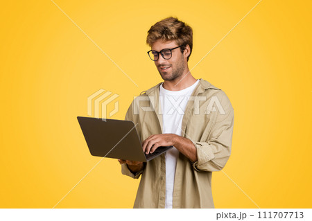 European man typing on laptop, standing against yellow background 111707713