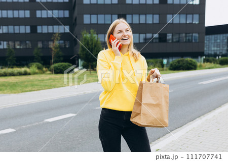 30s happy woman holding a paper bag in hands and calling on phone on city street. Technology, communication and people concept 111707741