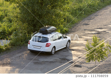New clean white modern car with black roof luggage box container moving along empty asphalt road in bad condition by green trees and bushes on bright sunny day New clean white modern car with black roof luggage box container moving along empty asphalt road in bad condition by green trees and bushes on bright sunny day 111708059