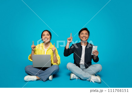 Joyful woman and man sitting with tech devices, pointing upwards, indicating a shared idea 111708151