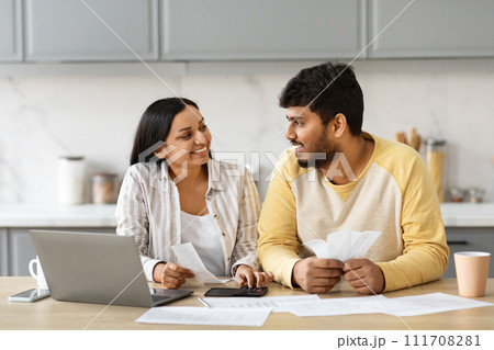 Smiling indian couple with laptop in kitchen calculating bills Smiling indian couple with laptop in kitchen calculating bills 111708281