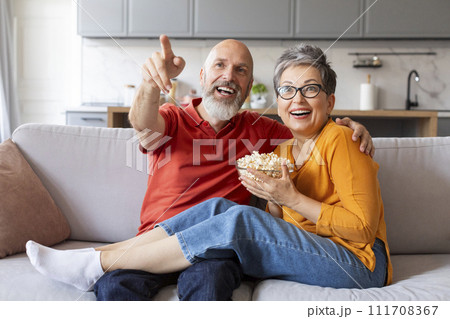 Cheerful senior spouses watching tv and eating popcorn at home Cheerful senior spouses watching tv and eating popcorn at home 111708367