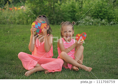 A comical happy laughing girl is sitting next to her autistic sister, covering her face with a heart made of puzzles A comical happy laughing girl is sitting next to her autistic sister, covering her face with a heart made of puzzles 111708759