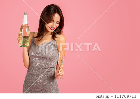 Joyful woman in a glittering silver dress holding a bottle of champagne and a glass 111709713