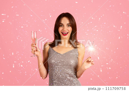 Surprised and joyful woman in a glittering silver dress holding a glass of champagne 111709758
