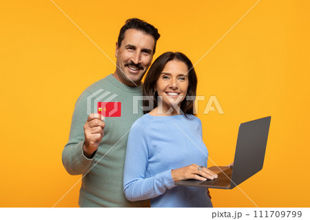 Cheerful couple engages in online shopping, man showing credit card, woman types on laptop Cheerful couple engages in online shopping, man showing credit card, woman types on laptop 111709799