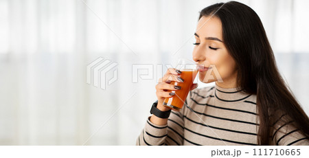Savoring the moment, a young woman in a striped turtleneck gently enjoys her carrot juice 111710665