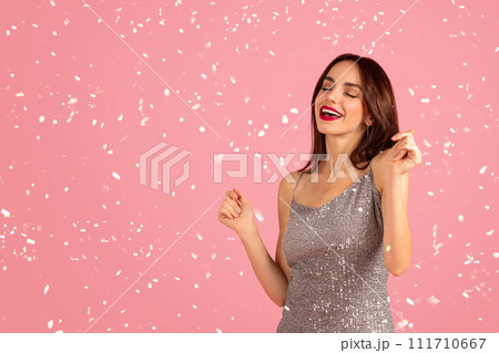 A joyous woman in a sequined silver dress dances with eyes closed and a smile 111710667