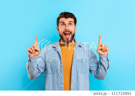 Positive young Caucasian man points upward over blue backdrop Positive young Caucasian man points upward over blue backdrop 111710991