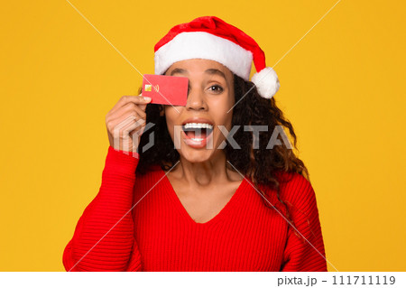 Excited black lady wearing Santa hat, hides eye with credit card, yellow background 111711119