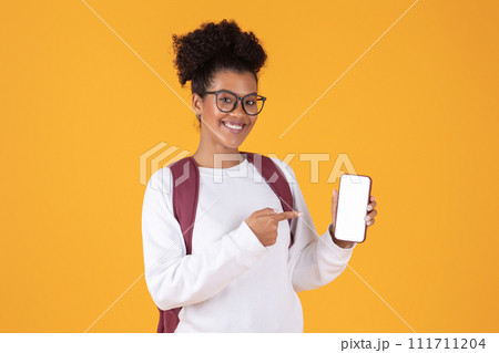 Happy young black woman wearing glasses and backpack pointing at blank smartphone 111711204