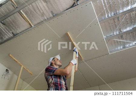 Young worker in protection work gloves fixing wooden holders for drywall suspended ceiling to metal frame insulated with shiny aluminum foil. DIY, do it yourself concept. Young worker in protection work gloves fixing wooden holders for drywall suspended ceiling to metal frame insulated with shiny aluminum foil. DIY, do it yourself concept. 111711504