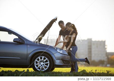 Young couple, handsome man and attractive woman at car with popped hood checking oil level in engine using dipstick on clear sky background. Transportation, vehicles problems and breakdowns concept. 111711560