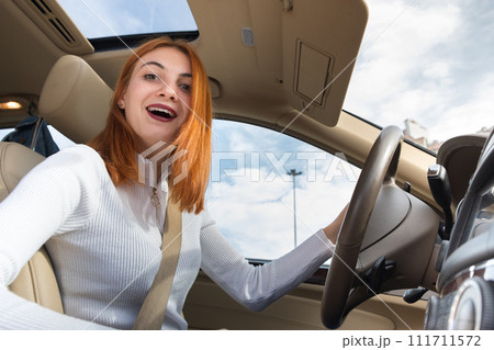 Wide angle view of young redhead woman driver fastened by seatbelt driving a car smiling happily. 111711572