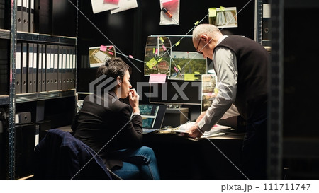 Team of detectives working to solve case, conducting criminal investigation to bring justice. Two police agents evaluating incriminating documents and clues to determine evidence authenticity. Team of detectives working to solve case, conducting criminal investigation to bring justice. Two police agents evaluating incriminating documents and clues to determine evidence authenticity. 111711747