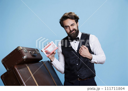 Hotel employee using heart pills to stop pain on camera, working in hospitality industry but dealing with cardio issues. Professional bellhop posing with package of pills and medicaments. 111711804