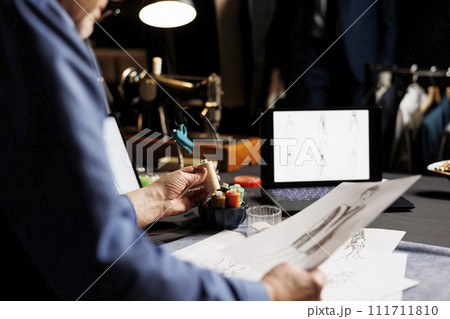 Close up shot of tailor picking sewing materials and sketch design inspiration for customer comissioned attire. Couturer in tailoring studio starting work on sartorial collection 111711810