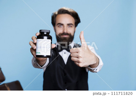 Bellhop recommends bottle of pills to help with immune system on camera, showing jar of vitamins for healthcare. Professional employee uses supplements in studio with blue background. 111711820