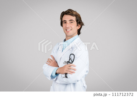 Smiling confident male therapist in white coat holding stethoscope, crossed arms on chest isolated on gray background 111712372