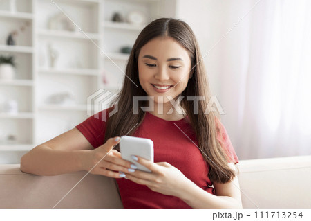Smiling young asian woman focused on texting with her smartphone at home 111713254