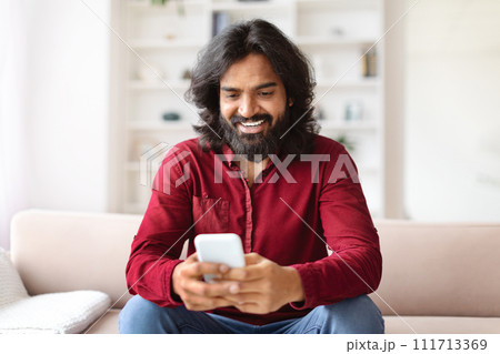 Happy Indian Guy Using Smartphone Browsing Internet Sitting On Couch Happy Indian Guy Using Smartphone Browsing Internet Sitting On Couch 111713369