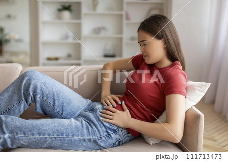Asian woman experiencing abdominal pain at home, holding her belly Asian woman experiencing abdominal pain at home, holding her belly 111713473
