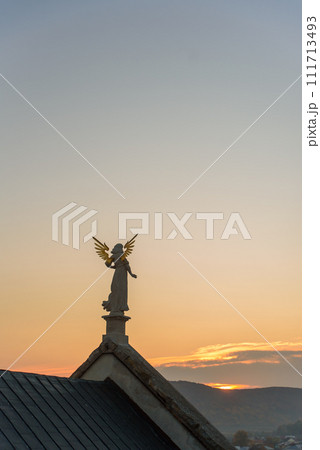Stone sculpture of an angel with golden wings standing on the rooftop of a baroque temple at sunset. 111713493