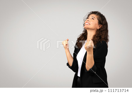 Elated young european businesswoman with beaming smile looks up, raising her fists in victorious gesture, free space 111713714