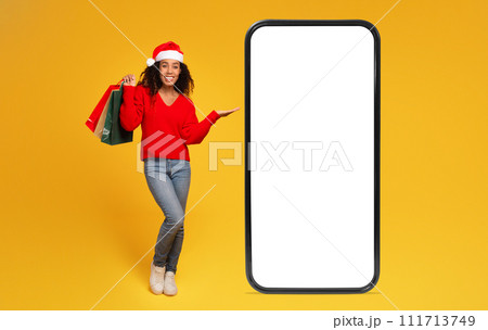 Black lady in Santa hat holds bags and shows phone with blank screen for sales, yellow background Black lady in Santa hat holds bags and shows phone with blank screen for sales, yellow background 111713749